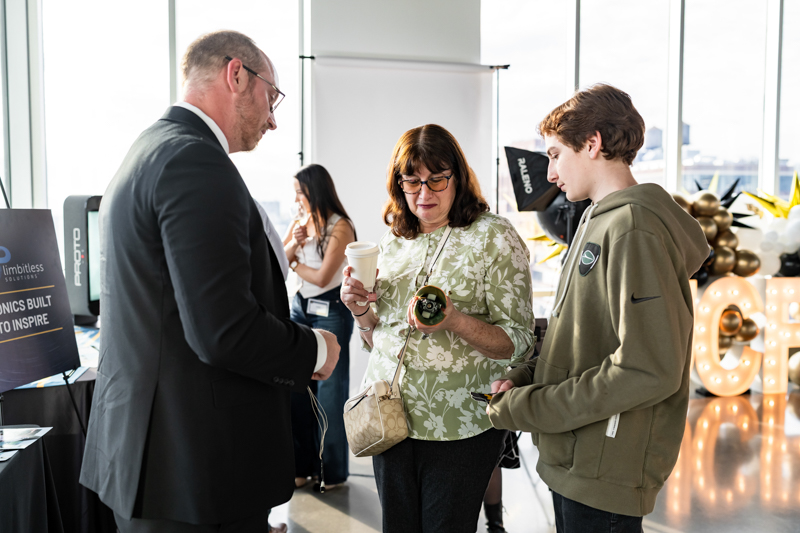 Professional man in a suit chats with a woman holding a coffee cup at a bright event booth, a young man stands nearby holding a badge lanyard.