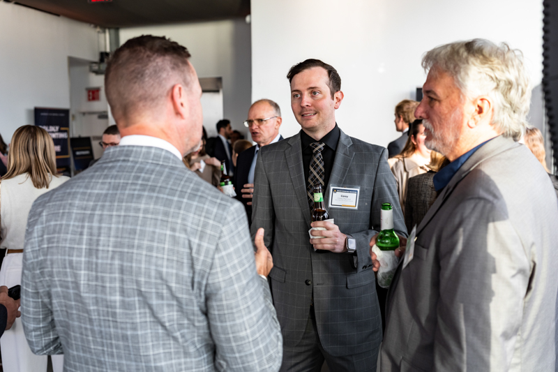 Business networking event: professionals in suits chat in a hallway, one man holding a beer bottle and name tag visible in the center.