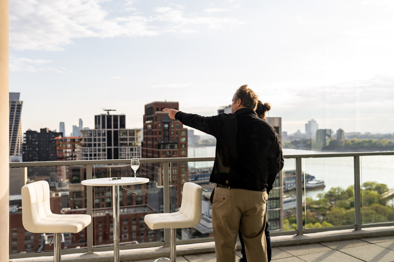 Two people on a rooftop terrace overlooking a city skyline, one gesturing toward the distance; a glass of wine on a table.