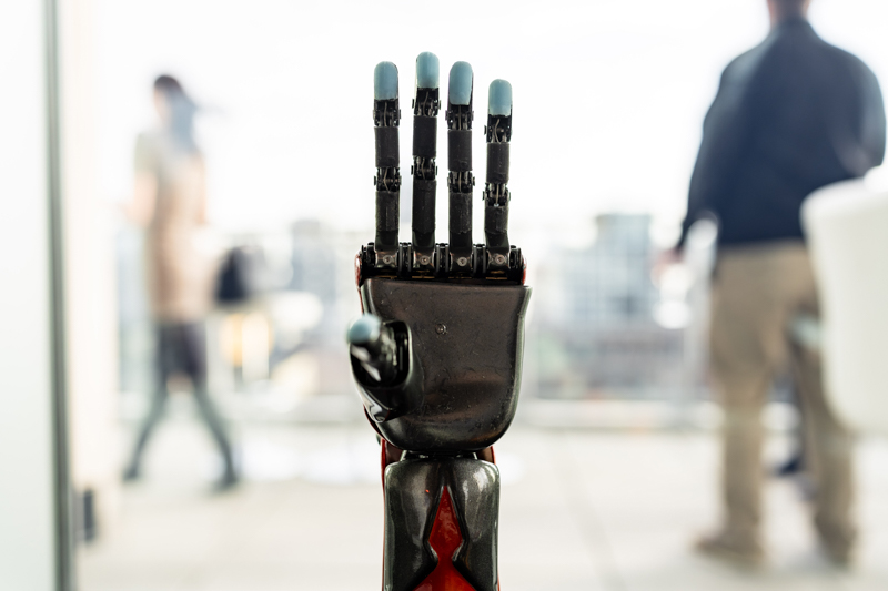 Robotic prosthetic hand with articulated fingers raised in a bright office setting with blurred coworkers in the background.