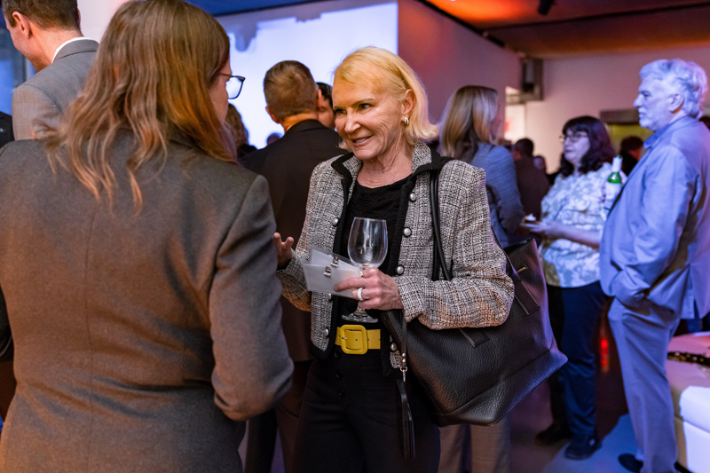 Business networking scene: a blonde woman in a tweed jacket chats with another attendee, holding a wine glass and papers.