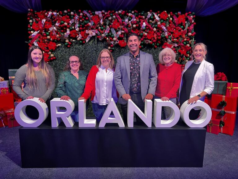 Six adults stand behind a large white 'ORLANDO' sign, with a festive Christmas backdrop of red poinsettias, ribbons, and gifts behind them.