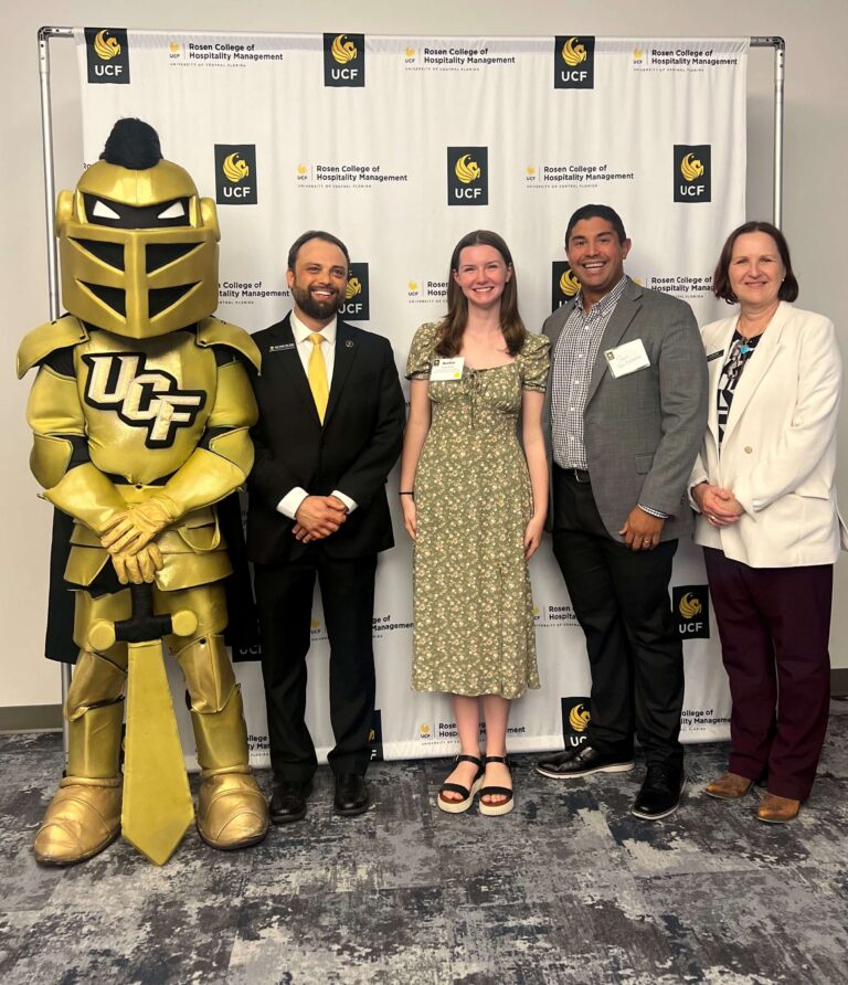 Five people stand with a gold knight mascot in front of a UCF Rosen College backdrop; smiles for a photo at an event.