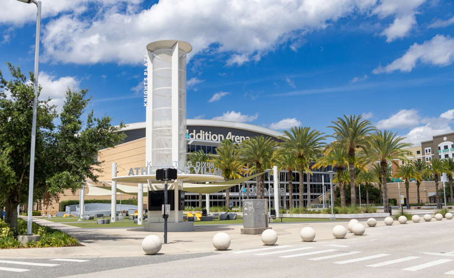 Exterior view of a large arena with a tall cylindrical sculpture, palm trees, and a row of spherical bollards along the street, under a blue sky.