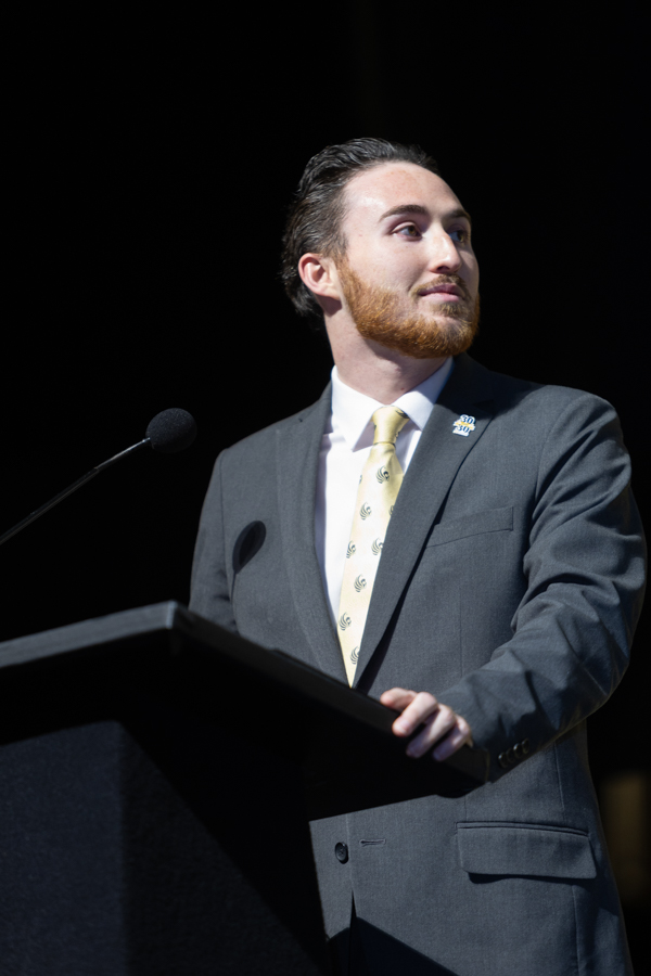 Man in a gray suit and yellow tie stands at a podium delivering a speech, looking to the right with a microphone nearby