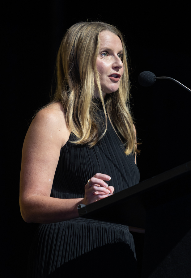 Woman in a black pleated dress speaking at a podium with a microphone on stage.