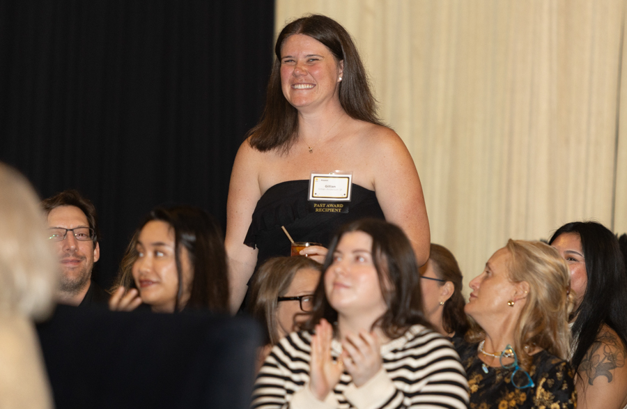 Woman in a black strapless dress stands on stage, smiling, with a name badge at an awards event (past award recipient).