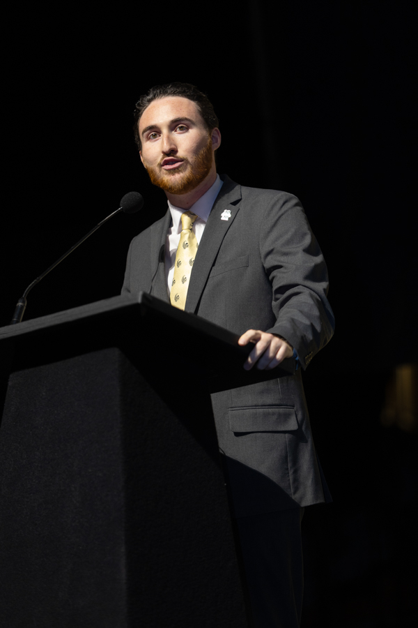 Man in a gray suit and yellow tie delivering a speech at a podium with a microphone