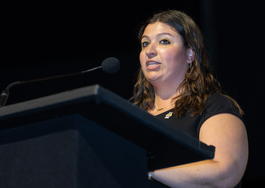 Woman with brown hair speaking at a podium with a microphone on a dimly lit stage in a black top/short sleeves.