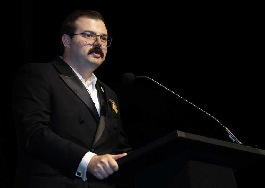 Man in a black tuxedo delivering a speech at a podium with a microphone on a dark stage