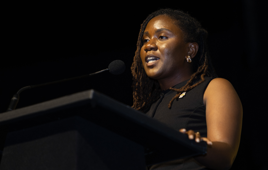 Black woman speaking at a podium with a microphone in a dark room, delivering a speech.