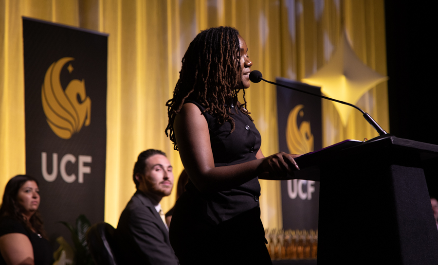 Woman speaking at a podium during a UCF event with banners in the background and seated audience members nearby.