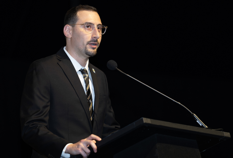 Man in a black suit and striped tie delivering a speech at a podium with a microphone on a dark stage.