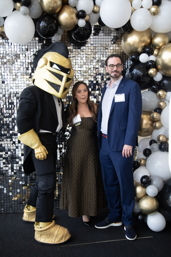 Three people posing for a photo at a festive event: a person in a gold knight mascot costume, a woman in a patterned dress, and a man in a blue suit with balloons behind them.