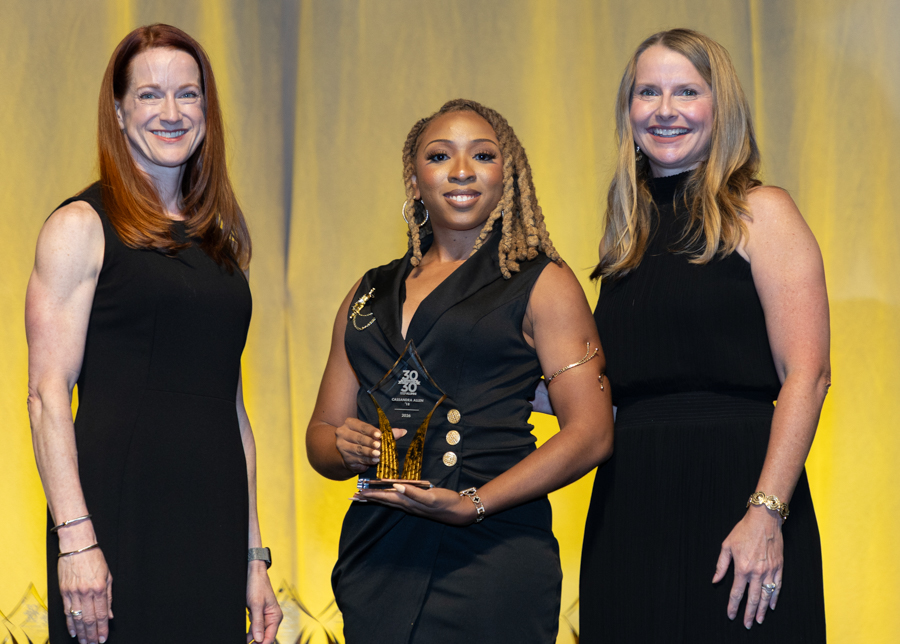 Three women in black dresses on a yellow-lit stage; center woman holds a glass trophy, flanked by two colleagues.