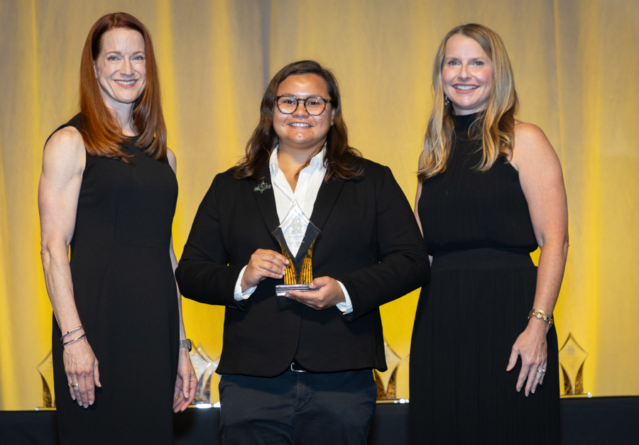 Three women in black attire on stage; center person holds an award trophy and smiles at the camera.