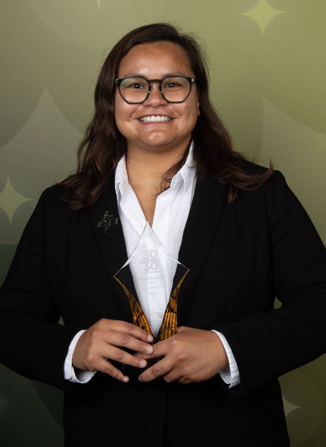 Professional woman in a black blazer and white shirt smiles while holding a glass trophy with a gold base in front of a green backdrop.