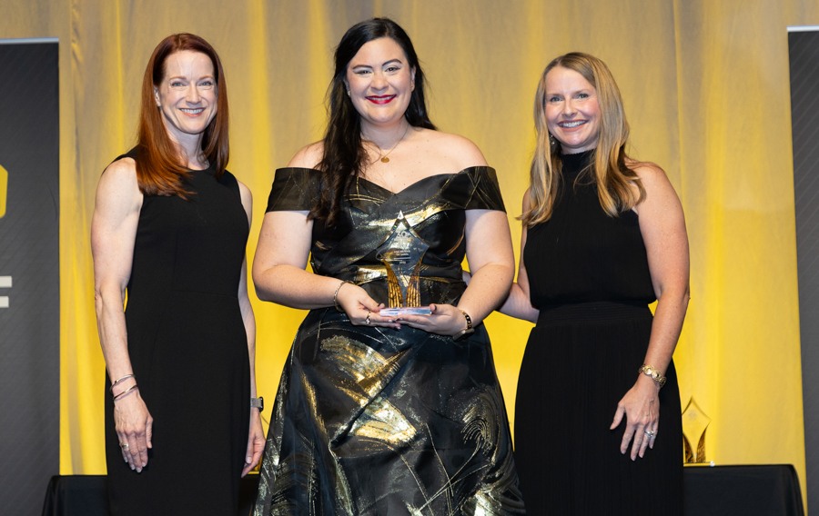 Three women in formal black dresses on stage, center holding a trophy with a yellow backdrop behind them.