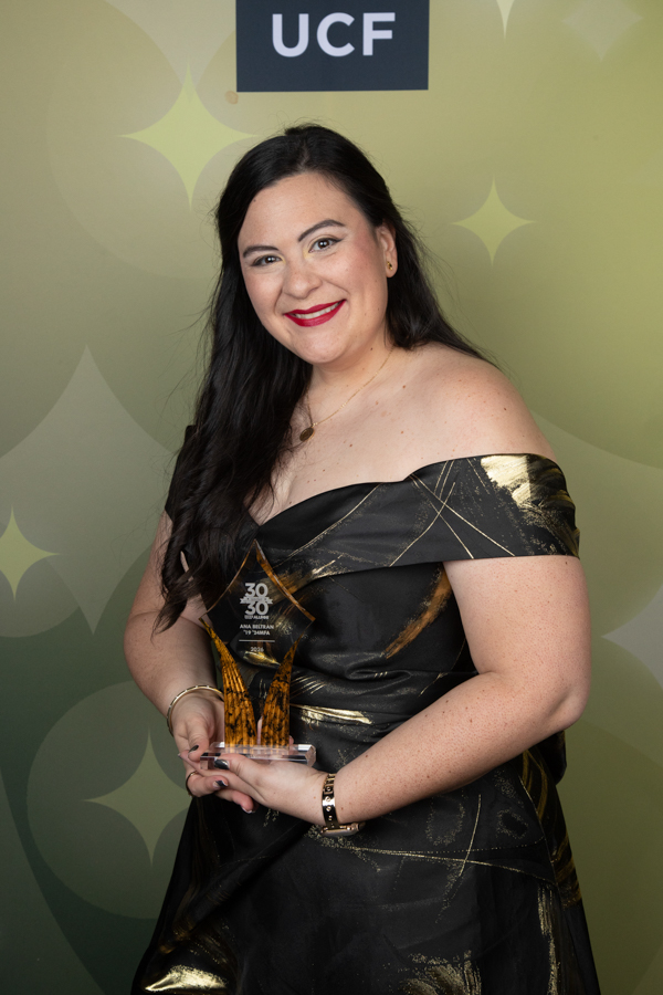 Smiling woman in an off-shoulder black and gold dress holds a crystal trophy at a UCF awards event, with a decorative green backdrop behind her.