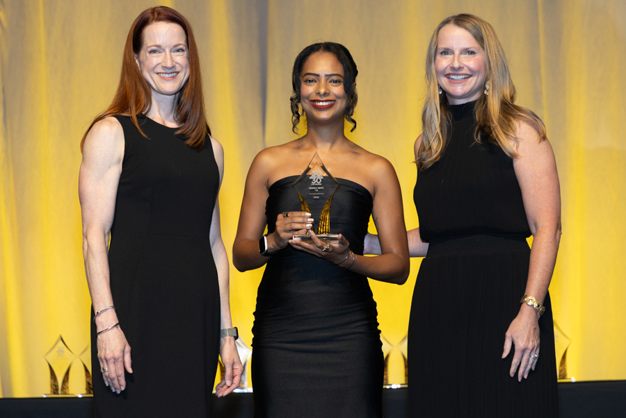 Three women in black gowns on stage; center woman holds a triangular glass trophy and smiles at the camera.