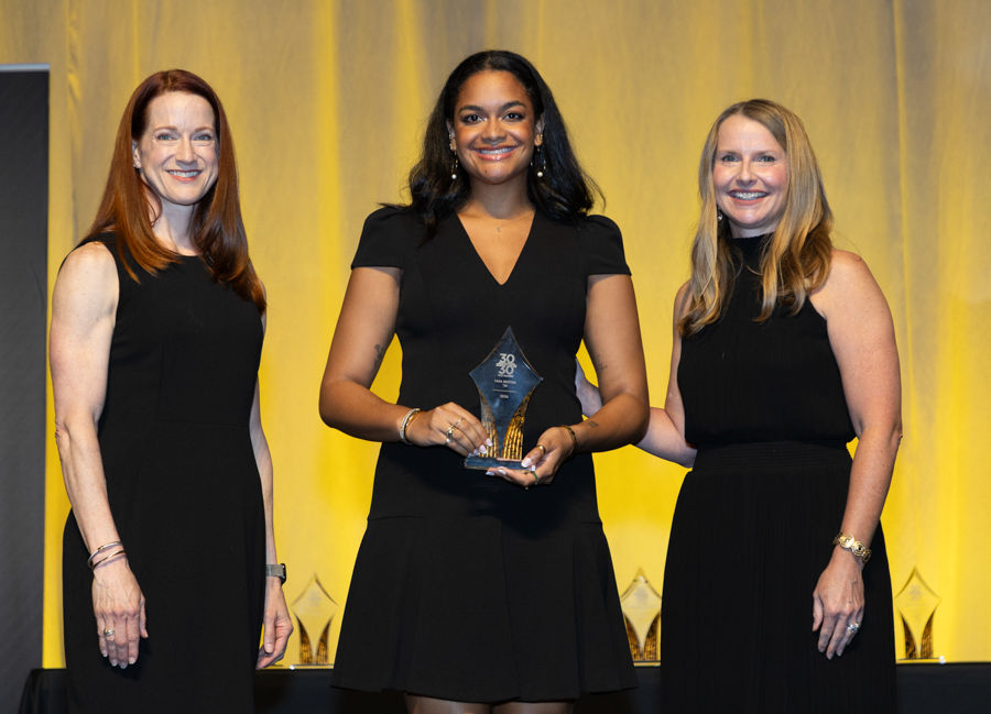Three women in black dresses on stage; the center woman holds a glass award and smiles toward the camera.
