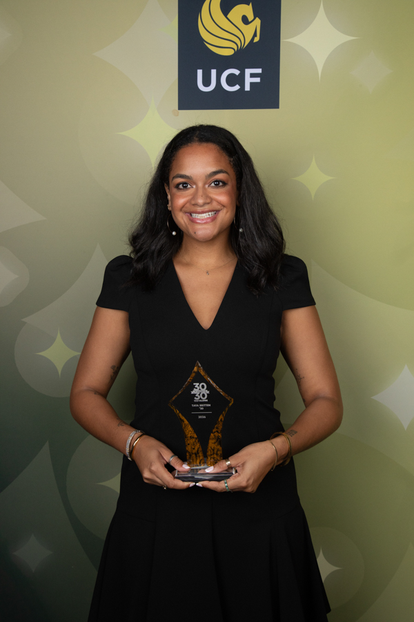 Smiling woman in a black dress holding a glass trophy, standing in front of a green backdrop with the UCF logo.