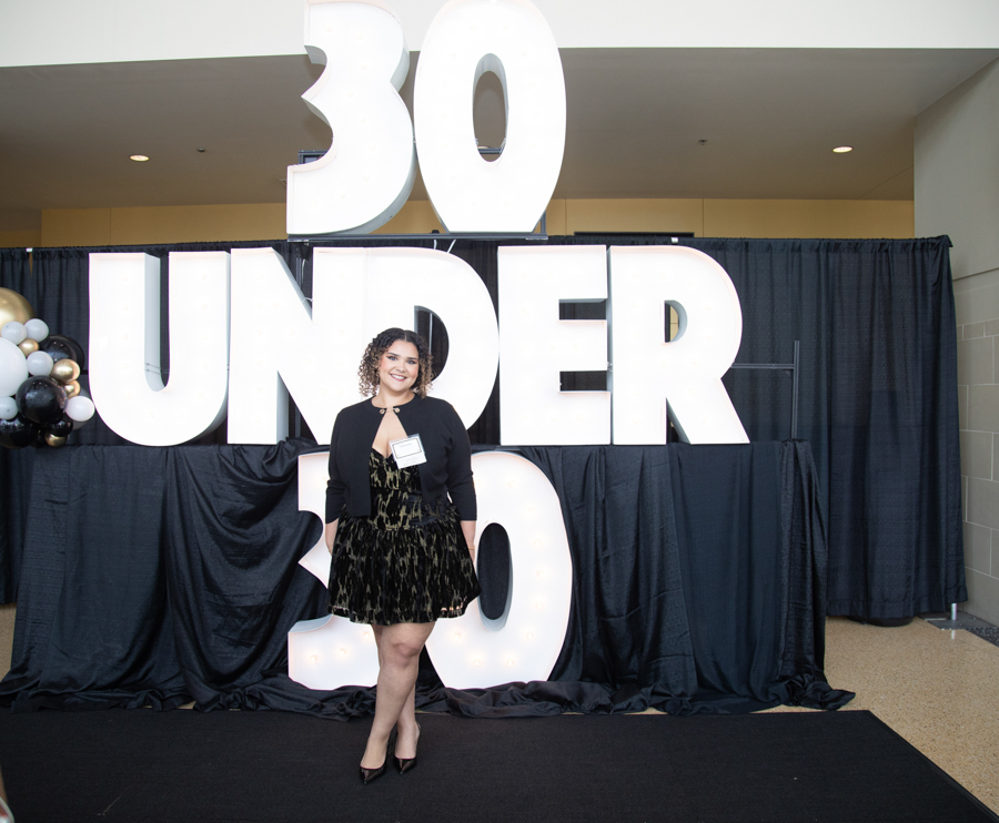 Woman in a black dress and blazer posing in front of a large '30 UNDER 30' backdrop at an event.