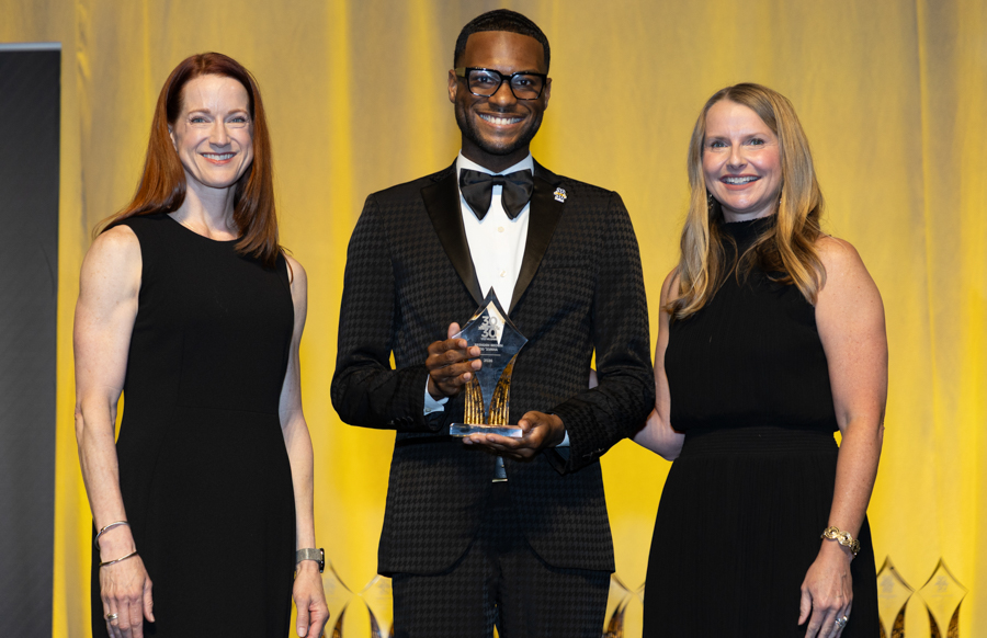 Three people pose on stage at an awards ceremony; man in a black tuxedo holds a glass trophy, flanked by two women in black dresses.