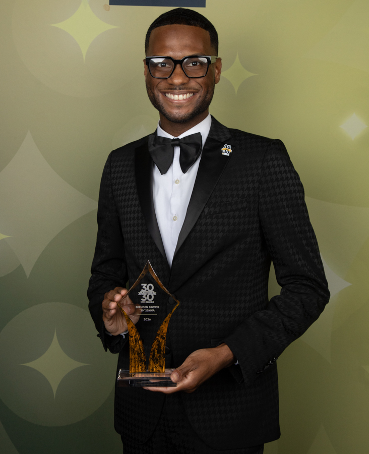Smiling man in a black checkered tuxedo and glasses holds a clear amber trophy at an awards ceremony with a green patterned backdrop.