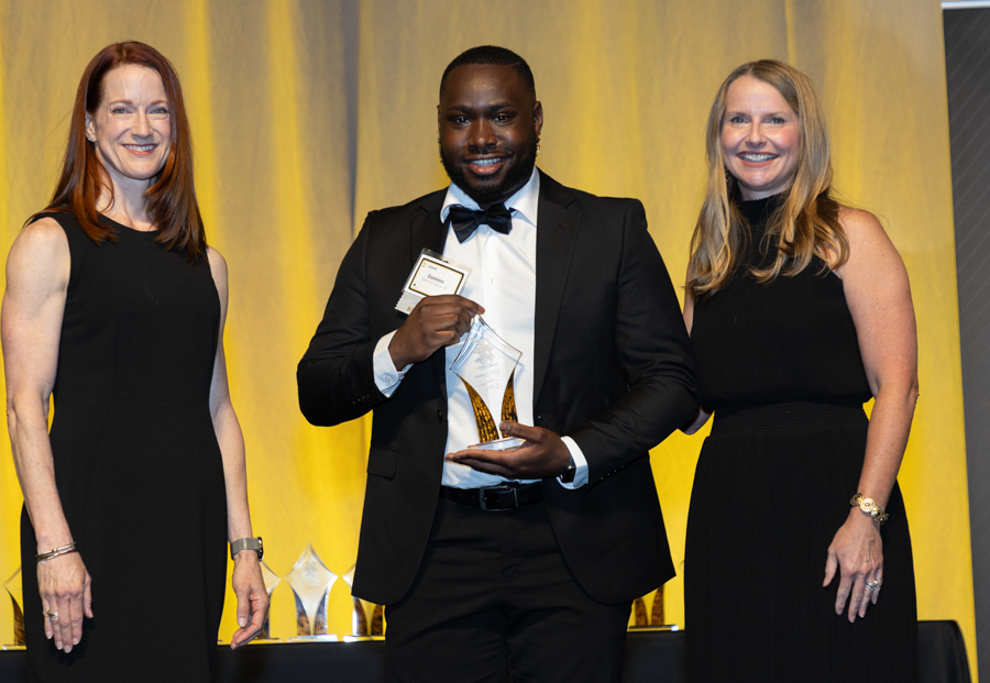 Man in a tuxedo holding a glass award on stage, flanked by two women in black dresses with a yellow backdrop.