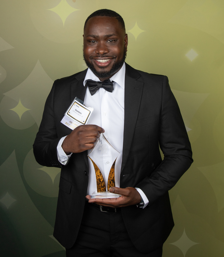 Smiling man in a black tuxedo holds a glass trophy with gold accents in front of a green patterned backdrop.