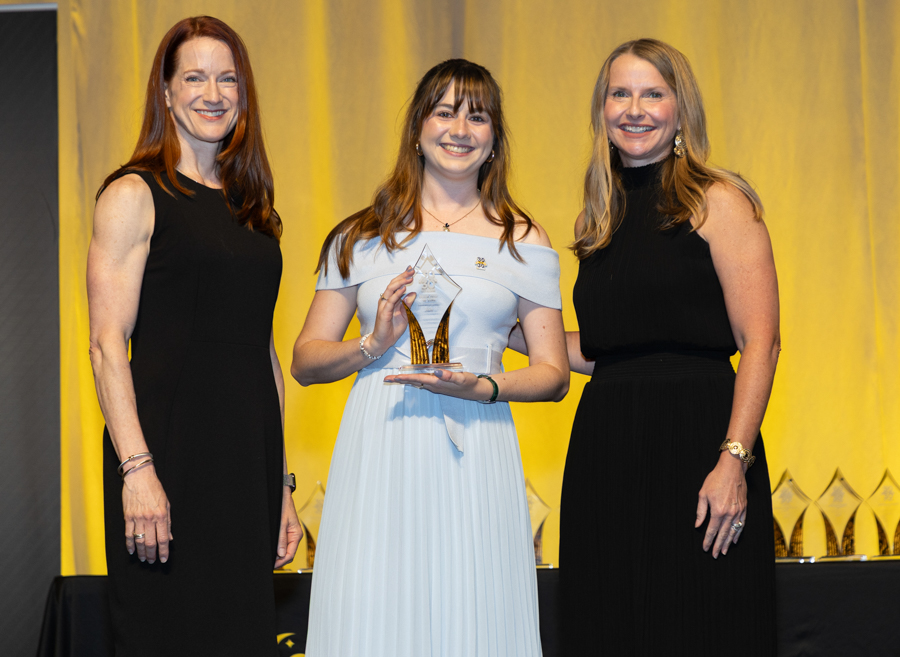 Three women in formal dresses stand on stage; the center woman holds a crystal trophy and smiles.