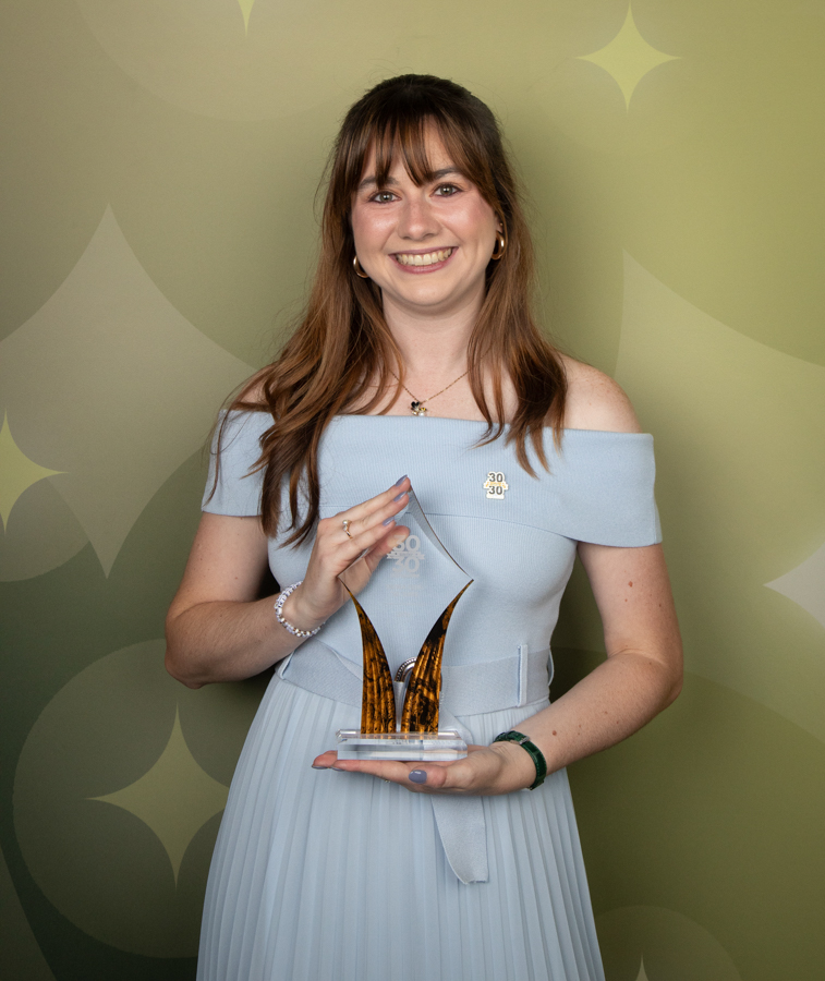 Woman in a pale blue off-the-shoulder dress holding a glass award with two tall amber spikes, smiling at the camera.