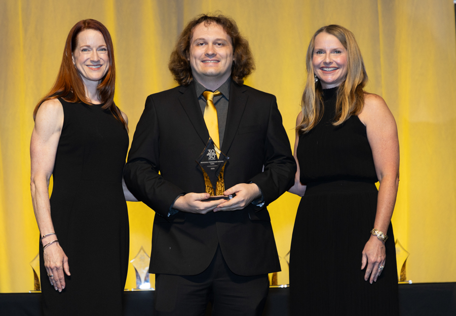 Three people in black formal attire on stage; center man holds a glass award, yellow curtain backdrop behind them.