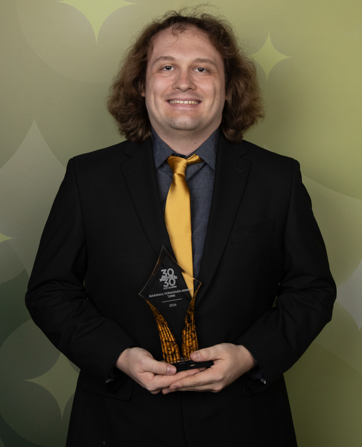 Smiling man in a black suit and yellow tie holding a glass diamond-shaped award against a green patterned backdrop.