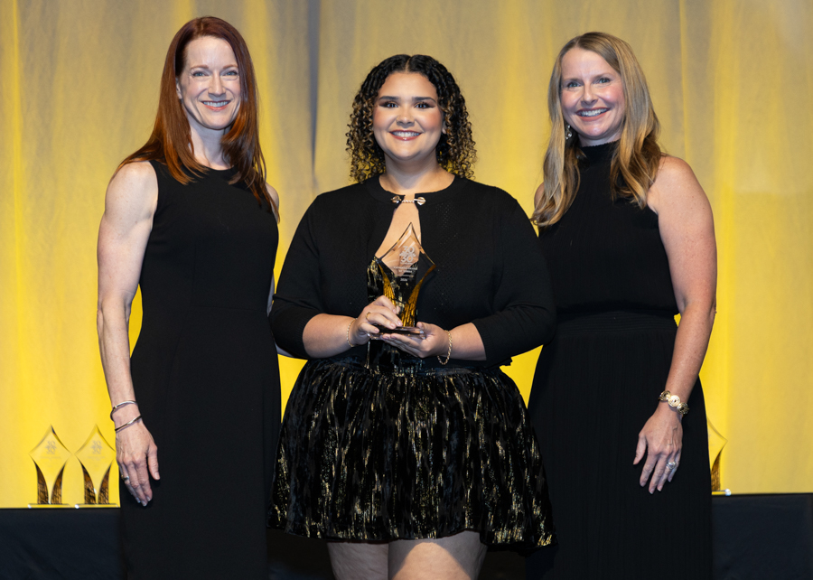 Three women in black formal dresses on stage; center holds a crystal award and smiles at the camera with a yellow backdrop behind them.