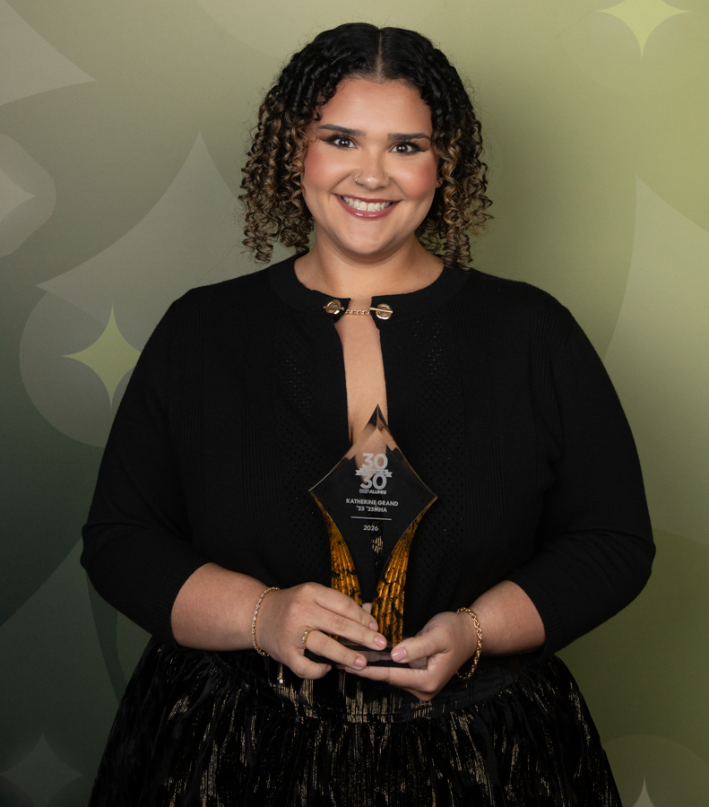 Smiling woman in a black outfit holding a glass award trophy in front of a patterned green backdrop.