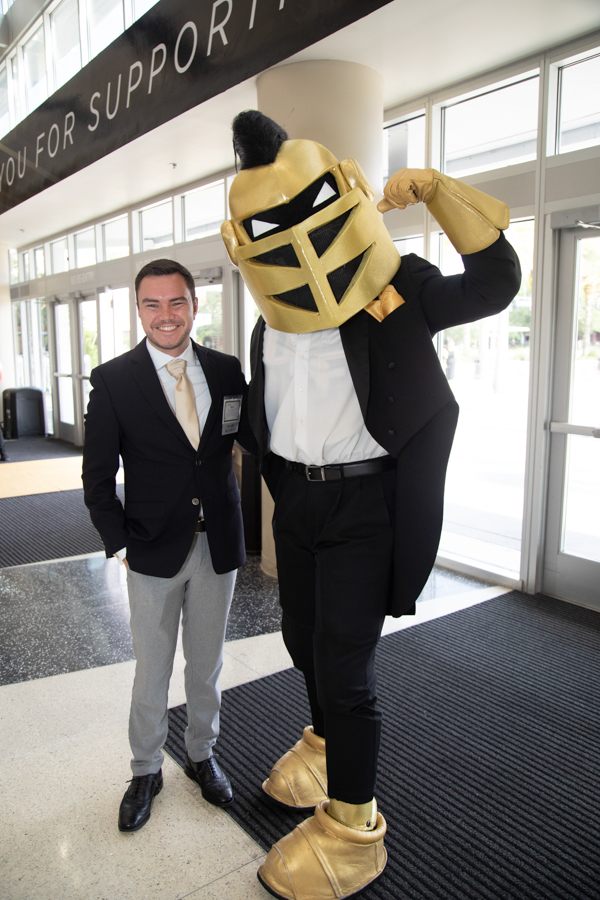 Smiling man in a blazer posing with a person in a gold knight mascot costume inside a lobby area with glass doors in the background.