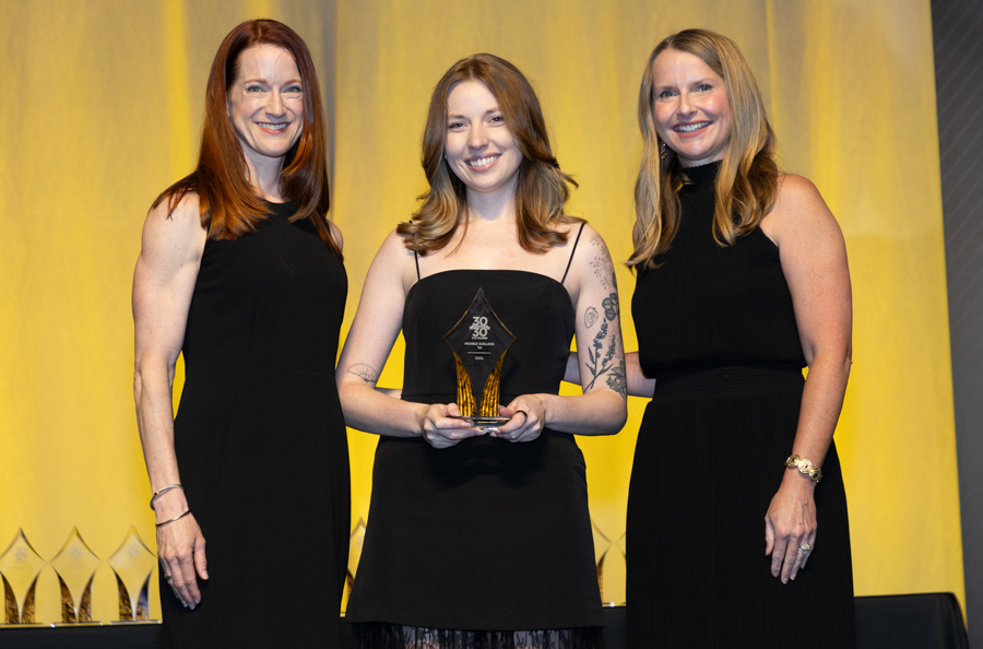 Three women in black dresses on stage; center woman holds a crystal trophy, yellow backdrop.