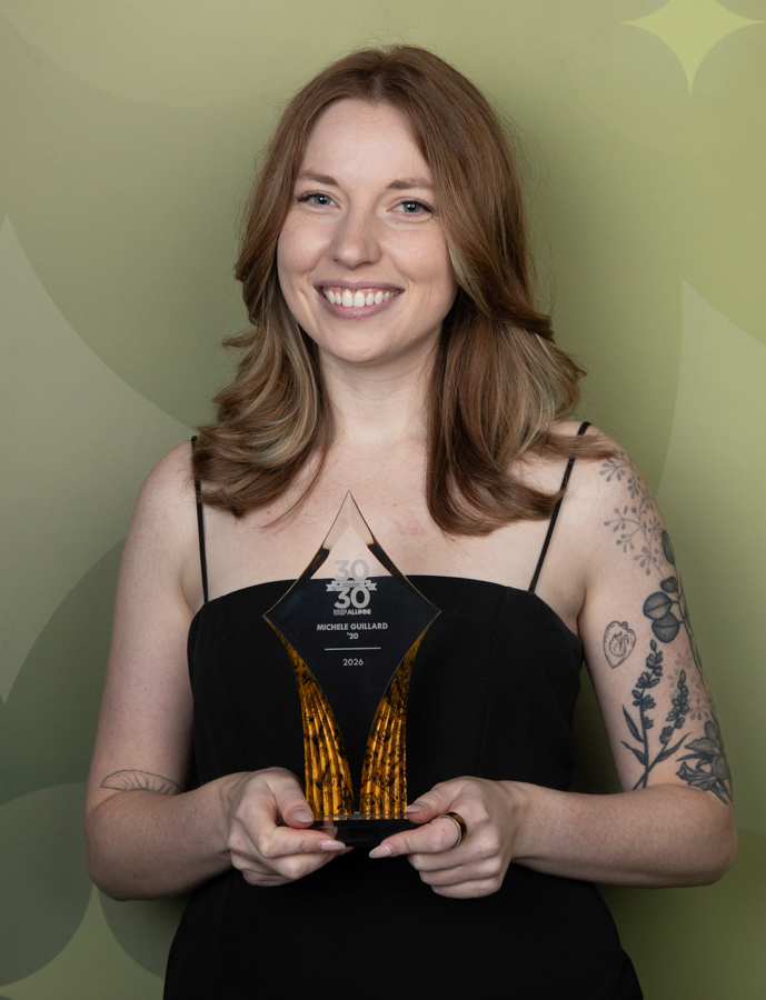 Smiling woman in a black dress holding a glass award with gold accents, inscription reads 30th anniversary and 2026, against a green pattern backdrop.