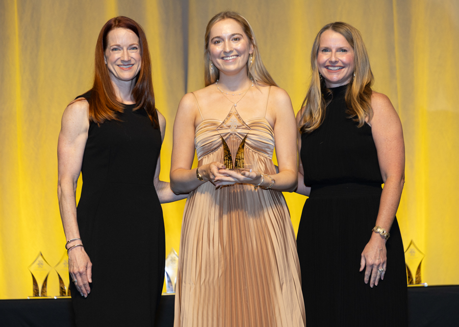 Three women on stage smiling; center woman in a peach gown holds a glass award, flanked by two women in black dresses.