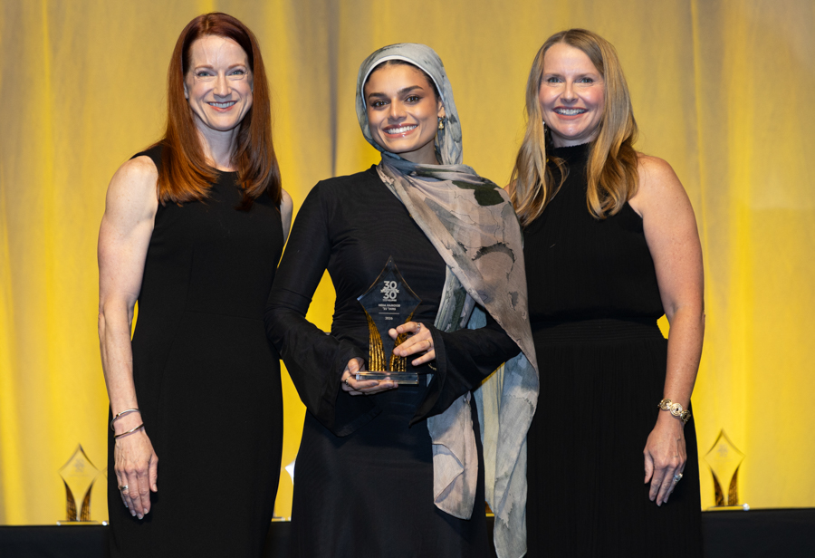 Three women in black dresses at an awards ceremony; the center woman wearing a hijab holds a glass trophy.