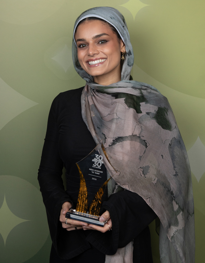 Smiling woman wearing a gray hijab holds a crystal award against a green patterned backdrop.