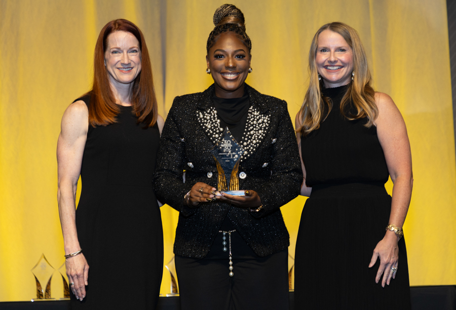 Three women in black dresses on stage; center woman in a glittery jacket holds an award trophy while smiling against a yellow backdrop. 2) Award ceremony with three women posing on stage; center figure wearing a sparkly jacket holding a glass trophy. 3) Group portrait at an awards event, featuring a smiling center figure in a jeweled blazer with a trophy. 4) On-stage photo of three women in black outfits; center recipient displays a glass trophy. 5) Award recipient flanked by two colleagues in black dresses at a ceremony, yellow backdrop.