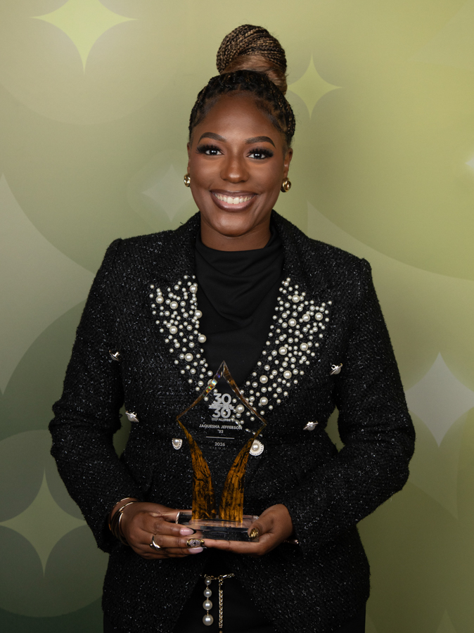 Smiling woman in a pearl-embellished black blazer holding a crystal award in front of a green backdrop.