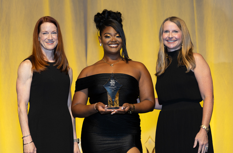 Three women in black evening gowns on a stage; the center woman holds a clear diamond-shaped trophy and smiles, flanked by two others against a yellow backdrop.