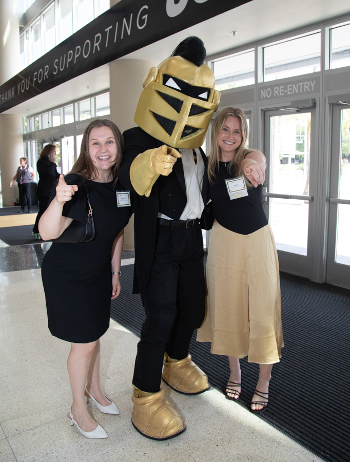 Two women with name badges pose and smile beside a gold armored knight mascot at an event entrance, thumbs up nearby.