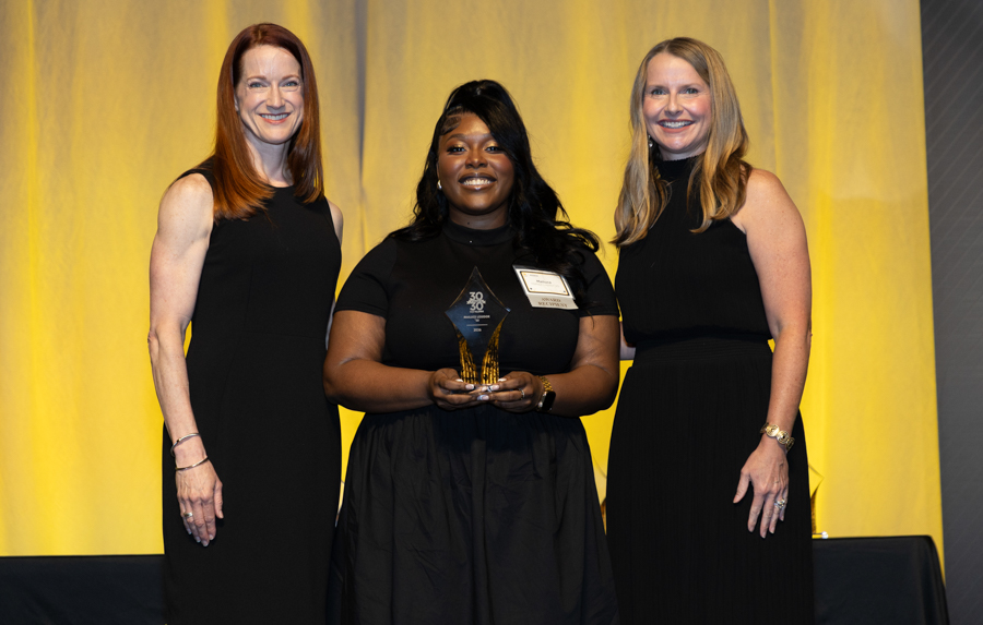 Three women in black dresses stand on a stage; the center woman holds a crystal award and smiles for the camera.