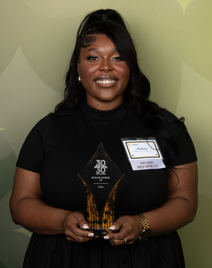 Smiling woman in a black top holds a tall glass award trophy and wears an 'Award Recipient' name badge at an awards event.