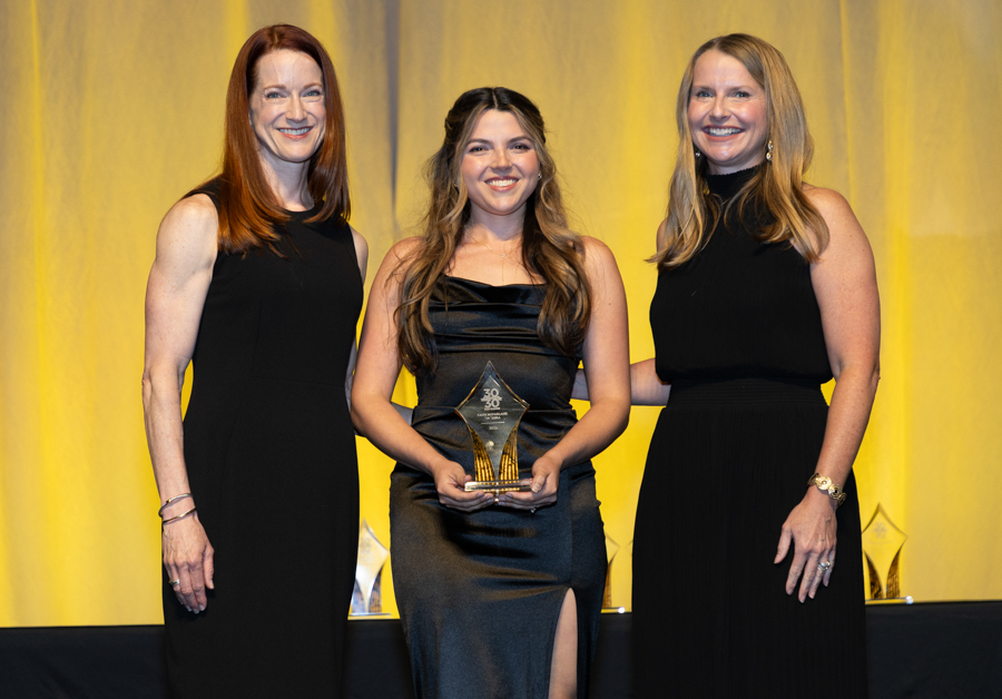 Three women in black evening gowns pose on a stage; the center woman holds a glass award and smiles at the camera.